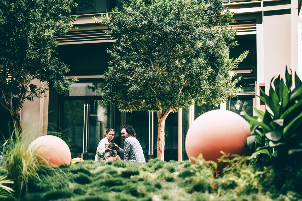 Two people meeting outdoors in a lush, modern courtyard with urban landscaping and sculptural elements. A contemporary scene of casual business or creative discussion surrounded by sustainable architecture and greenery. Conceptual image for eco-conscious work culture, urban innovation with biophilic design, and modern professional lifestyle.